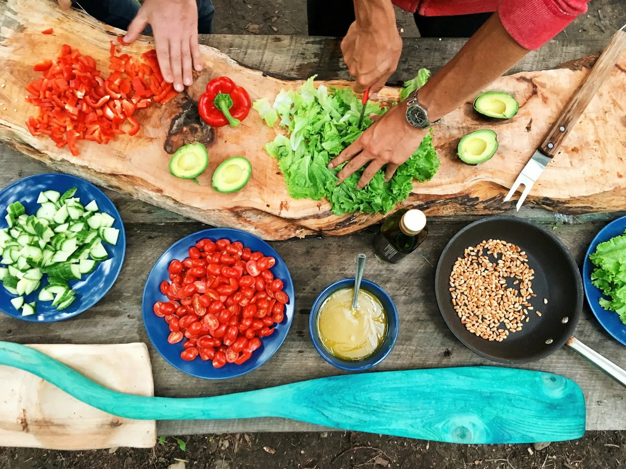 Hands chop red peppers and lettuce on a wooden board surrounded by bowls of cut vegetables