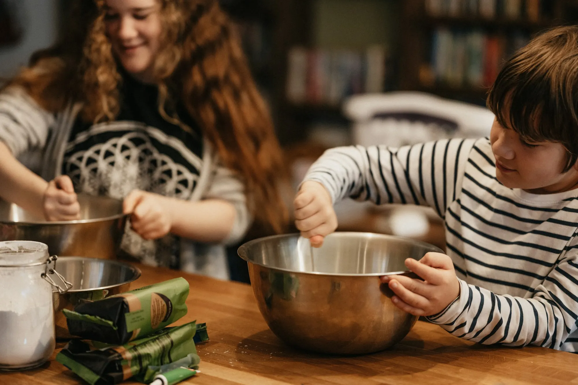 Two children mix ingredients in steel bowls at a wooden kitchen counter