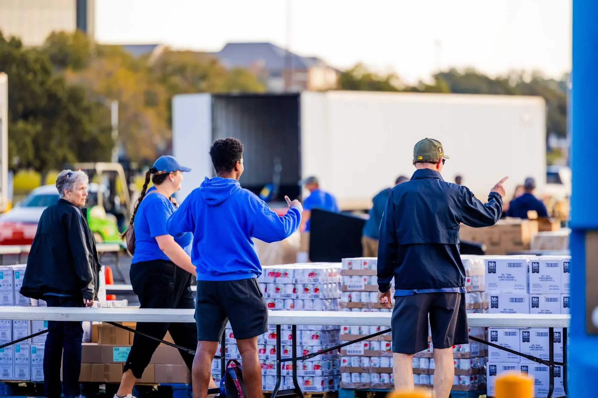 Volunteers at an outdoor food distribution, with tables and pallets of canned goods and a delivery truck behind