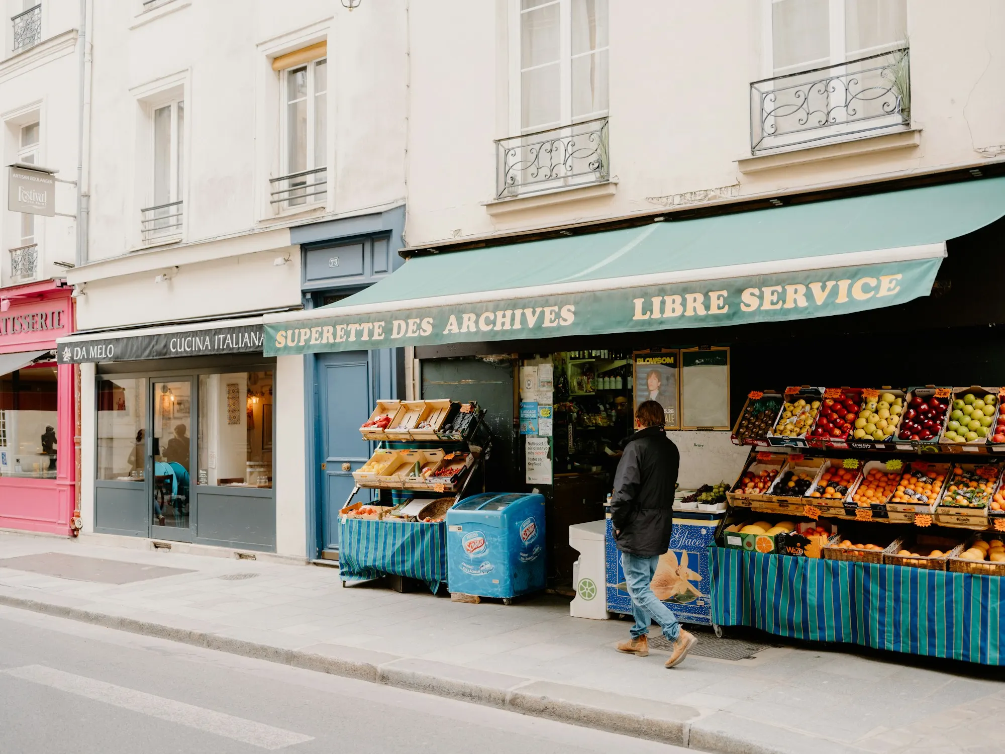 A man walks past a small corner grocery with fruit and vegetable stands on a quiet city street